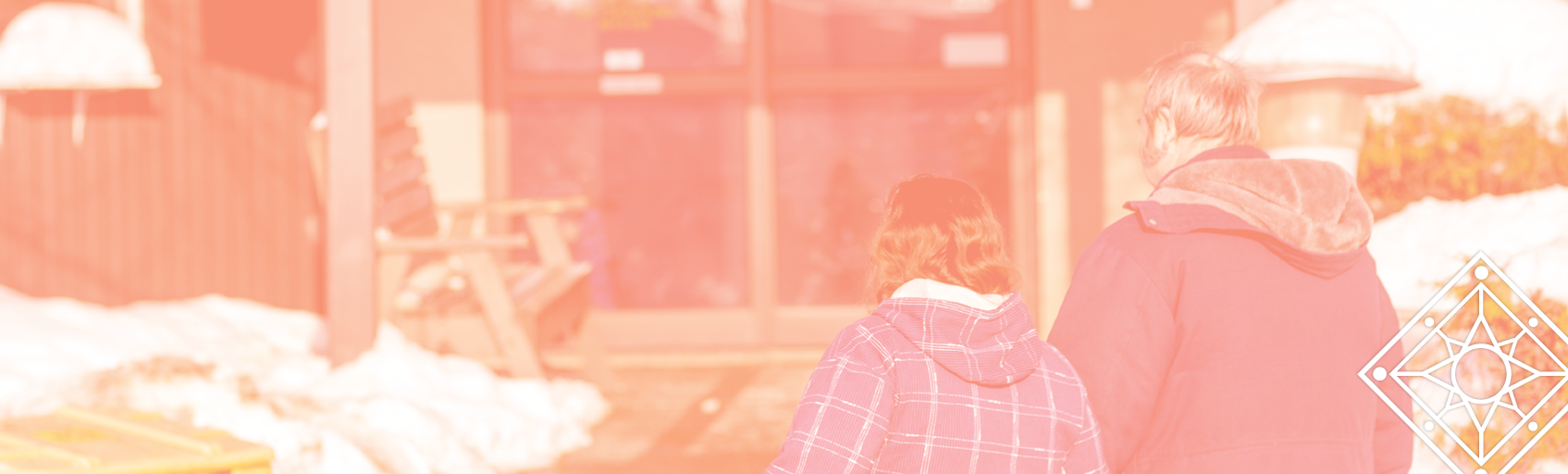 Two people outside in the daytime, wearing winter coats, walking toward the entrance of an apartment building with snow on the ground around them.