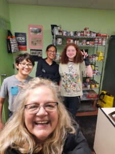 One adult and three youth in a green room, standing in front of a metal kitchen rack filled with groceries. The child on the right is making the peace gesture with her