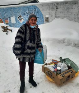 An adult in a knit poncho stands outside smiling in the snow with a large box of groceries at her feet
